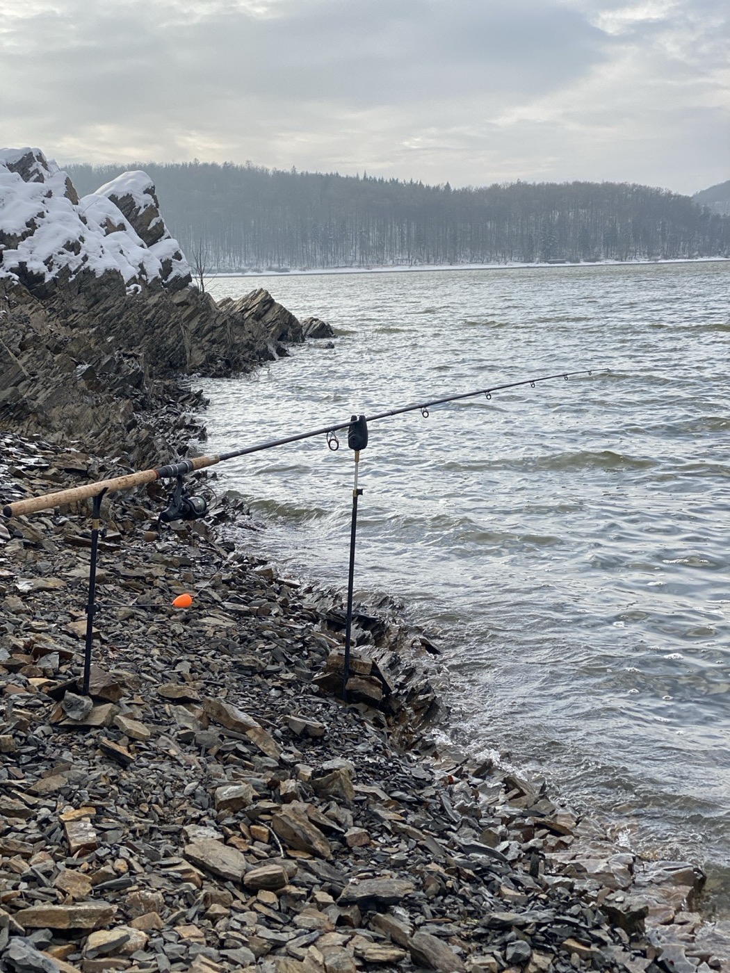 Bankstick mit Rute auf steinigem Ufer im Winter, Schnee auf den Felsen