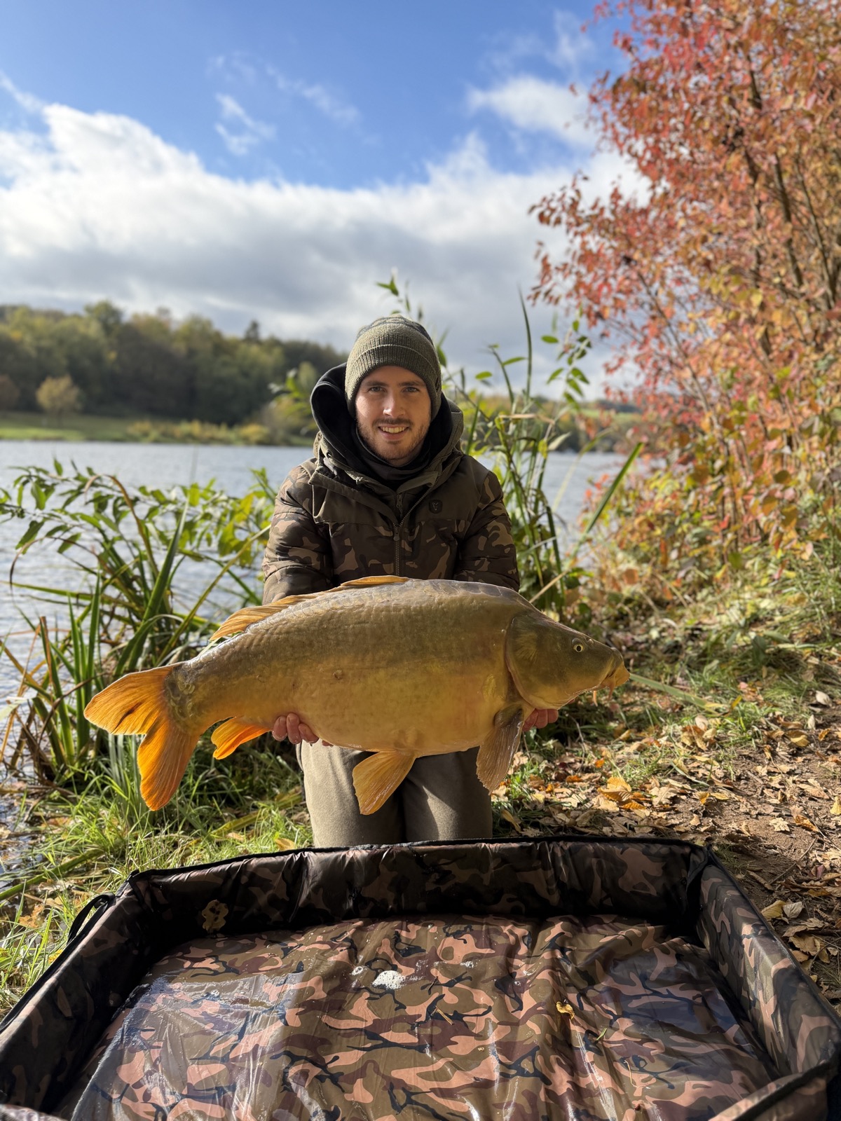 Fabi mit einem großen Spiegelkarpfen am Ufer, Herbstlaub im Hintergrund